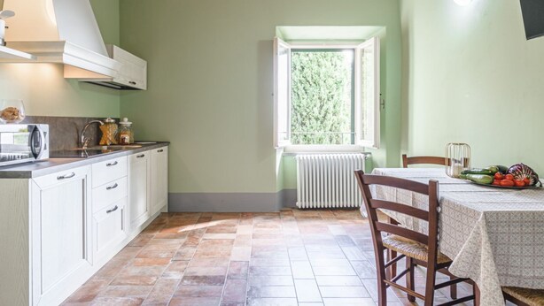 A kitchen with white cabinets, a window with a view of greenery, and a table with a tablecloth and vegetables. - Appartamento delle Logge (Cortona)