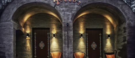 A stone building with two arched doorways, each with a light fixture and a chair in front.
