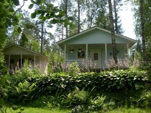 A two-story house with a porch, surrounded by dense greenery and tall trees. - Cabin-Garden view-Private Bathroom-Cottage-Mökki 11,The Middle Green (Västerskog)