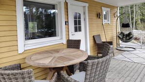 A covered patio with a round wooden table and wicker chairs, a hanging chair, and a door with a window. - Cabin-Garden view-Private Bathroom-Deluxe-Mökki 12,The Biggest Yell (Västerskog)