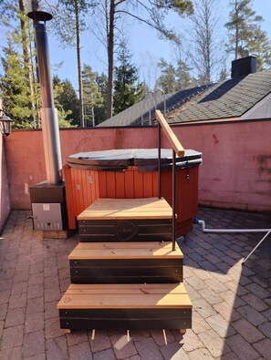 Boblebad med træterrasse og metalgelændere, foran en flisegul terrasse med røde vægge og træer i baggrunden.