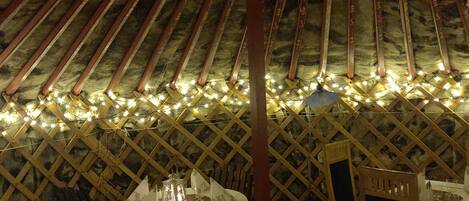 An indoor dining area with a thatched roof, wooden lattice walls, and string lights. Tables are set with white tablecloths and candles.
