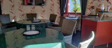 Un comedor con mesas redondas, sillas de mimbre, un televisor de pantalla plana y una zona de cocina con una encimera roja y varios objetos en estantes.