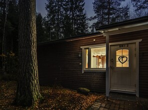Une cabane avec une façade en bois, une porte blanche portant l’inscription « BM12 » et une fenêtre ornée d’un motif en forme de cœur.