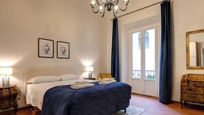 Une chambre à coucher avec un lit, deux lampes de chevet, un lustre, une commode et une fenêtre avec des rideaux.