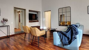 Un salon moderne avec un canapé, deux fauteuils, une table basse et un téléviseur. Un miroir est accroché au mur et une porte donne accès à une autre pièce.