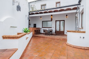 A courtyard with a tiled floor, a dining area with chairs and a table, and a white building with a tiled roof.