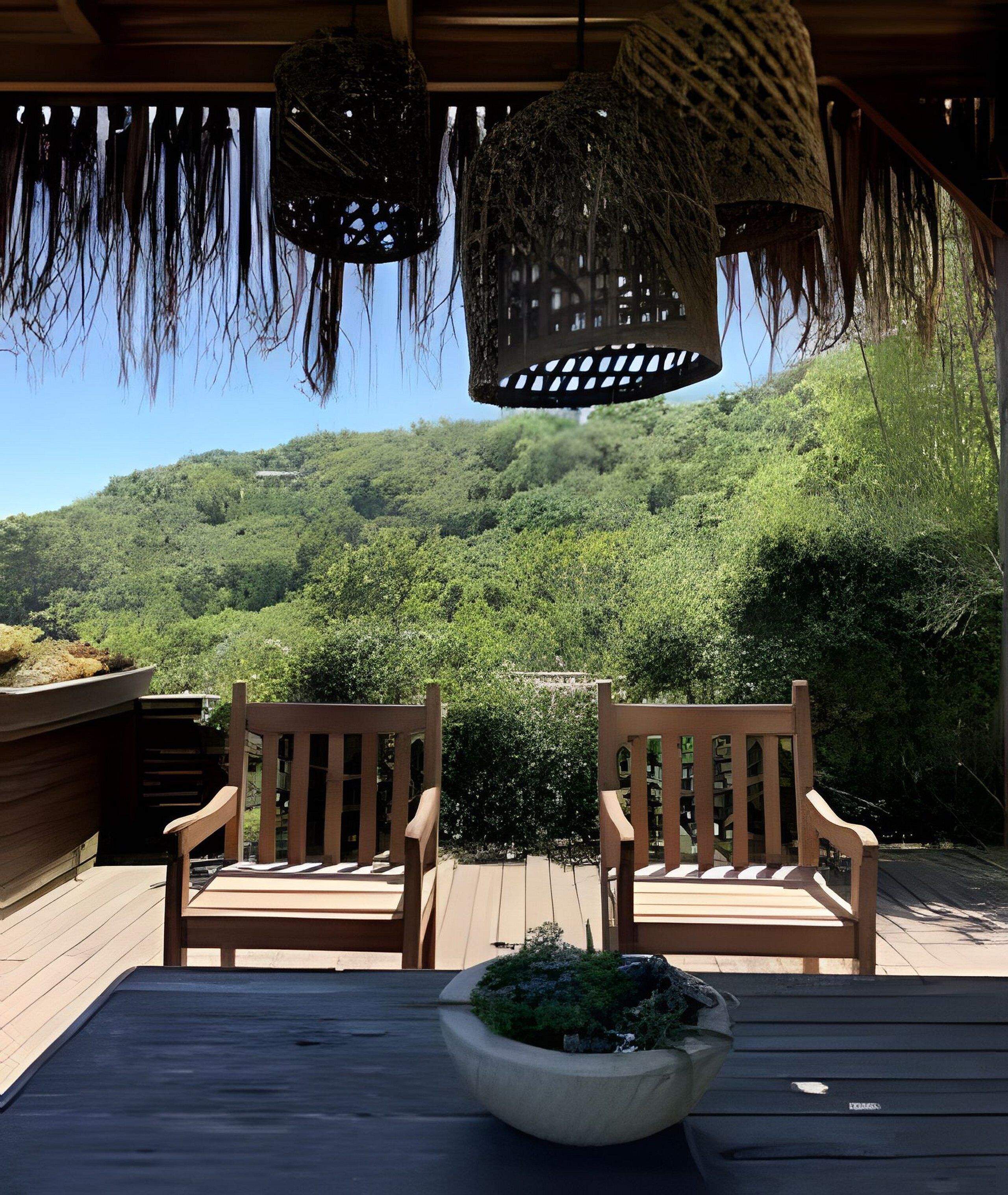 Uma área de estar ao ar livre com duas cadeiras de madeira e uma mesa, com vista para uma encosta arborizada.