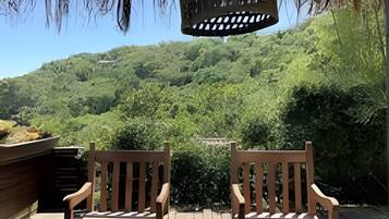 Uma área de estar ao ar livre com duas cadeiras de madeira e uma mesa, com vista para uma encosta arborizada.