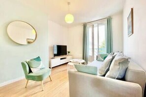 Een moderne woonkamer met een bank, een fauteuil, een televisie en een groot raam met gordijnen.