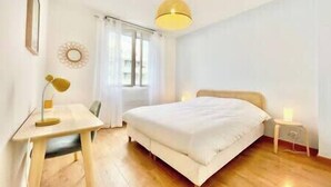 A bedroom with a bed, a desk, a chair, a bedside table, and a lamp.
