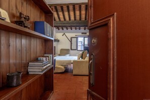 A narrow hallway leading to a bedroom with a bed, a chair, and a shelf with books and a lamp.