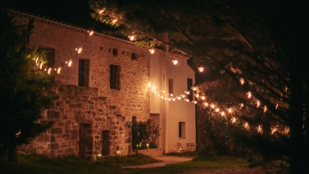 Un bâtiment en pierre décoré de guirlandes lumineuses, un sentier et un petit espace extérieur aménagé avec des sièges.