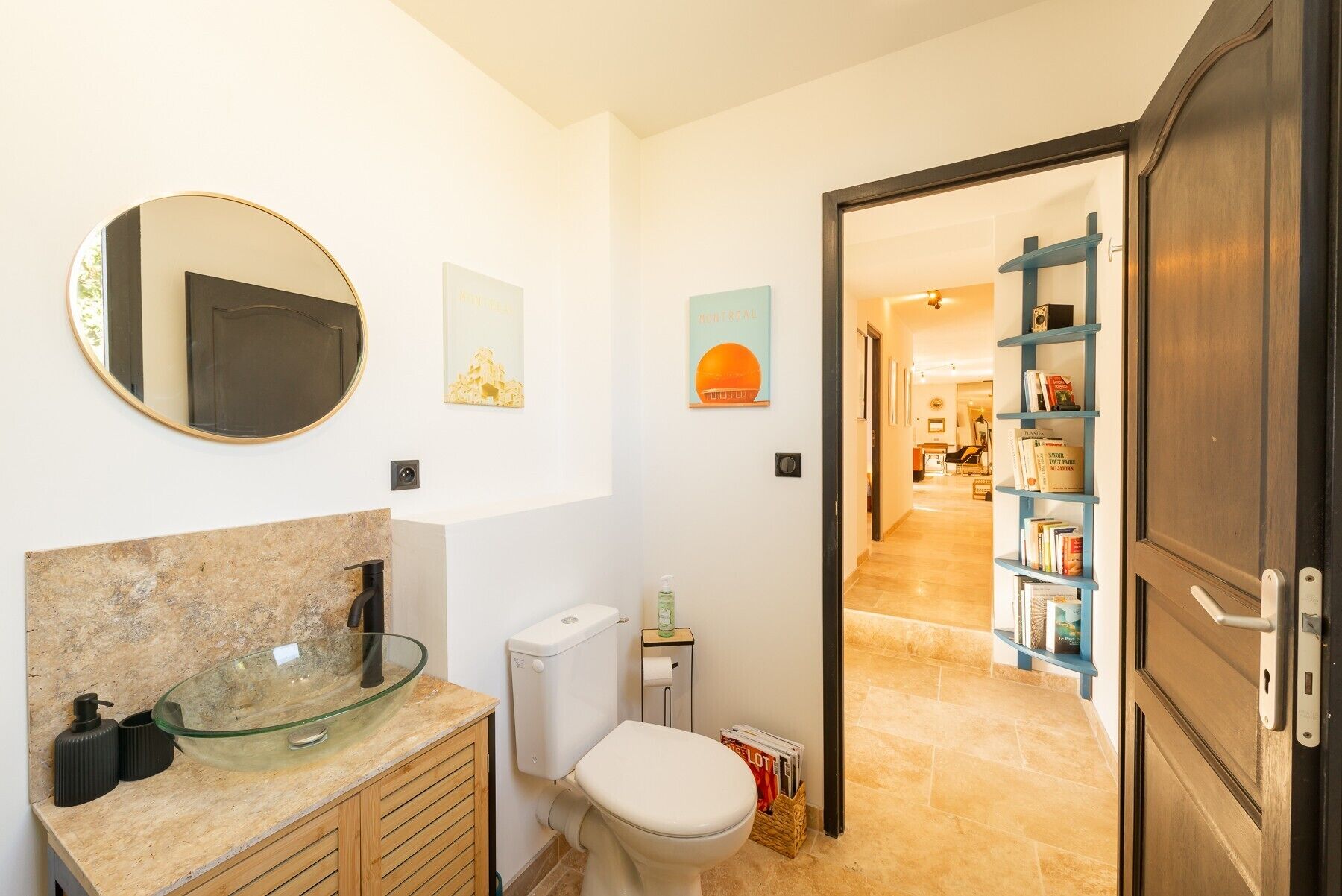 Un baño con un lavabo de vidrio, un inodoro y un espejo. Se ve una puerta que da a un pasillo con una estantería y un comedor.