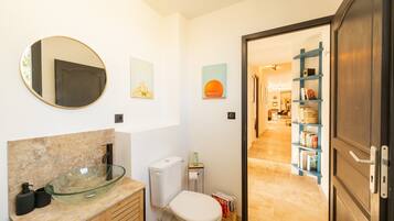 Un baño con un lavabo de vidrio, un inodoro y un espejo. Se ve una puerta que da a un pasillo con una estantería y un comedor.
