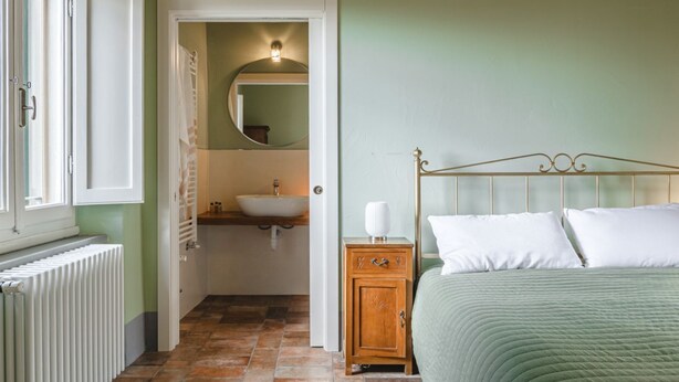 Ein Schlafzimmer mit einem Bett, einem Nachttisch, einer Kommode, einem Fenster und einem durch eine offene Tür einsehbaren Badezimmer.