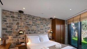 Une chambre moderne avec un mur en pierre, un grand lit recouvert de linge de maison blanc, deux lampes de chevet et une vue sur la verdure grâce à de grandes fenêtres.