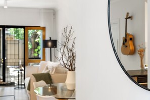 Un salon moderne avec une table basse en verre, un canapé et une lampe. On peut apercevoir une guitare dans le reflet du miroir.