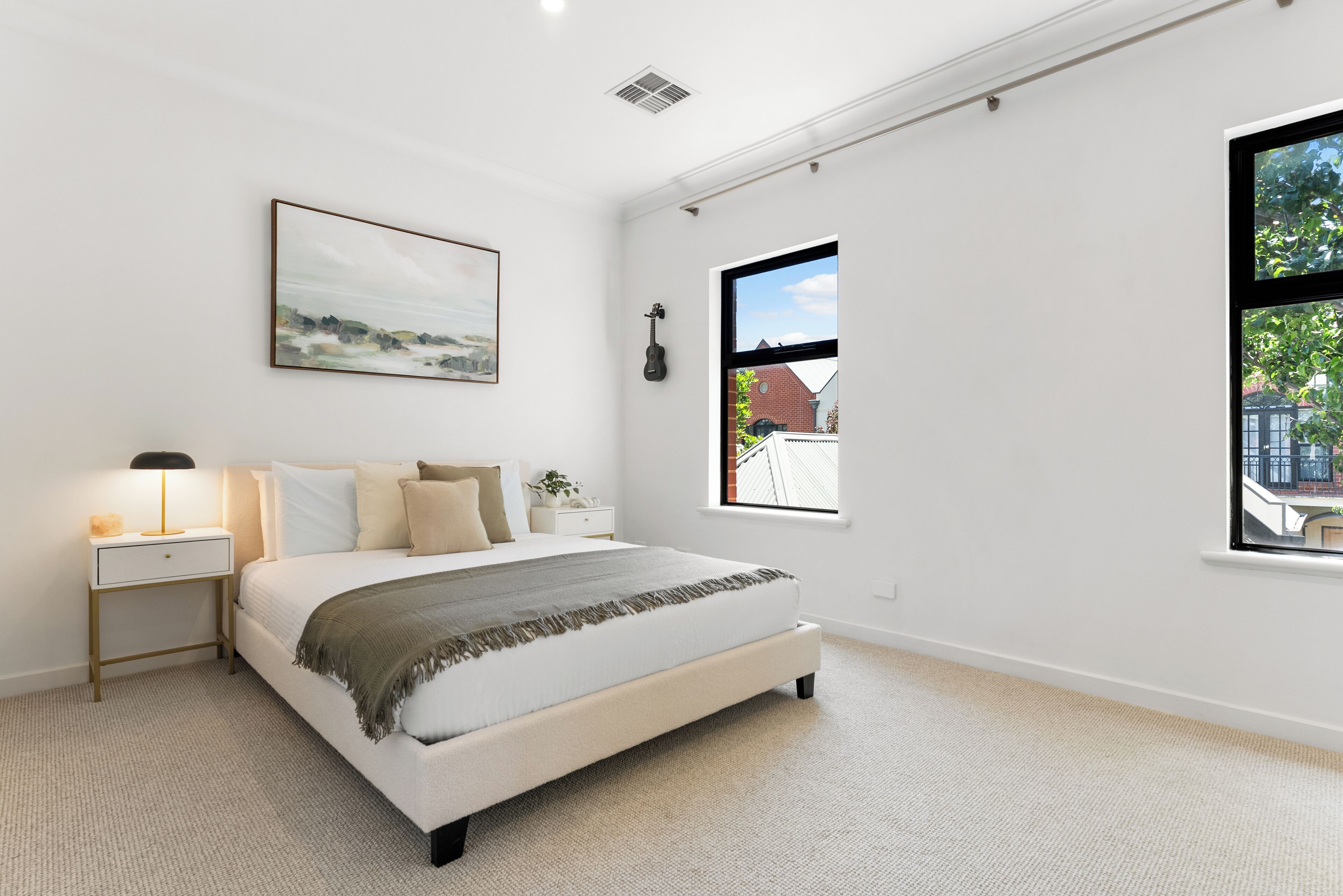 A modern bedroom with a large bed, a nightstand, a lamp, a framed picture, and a window with a view of buildings.