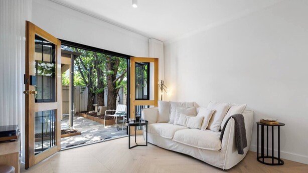 Un salon moderne avec un canapé blanc, une petite table d’appoint et une grande porte vitrée donnant sur un espace extérieur doté d’une terrasse en bois et de chaises.