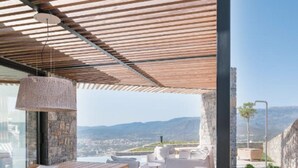 Uma área de refeições moderna ao ar livre, com mesa e cadeiras de madeira, luminária pendente e vista para a cidade e montanhas ao fundo.