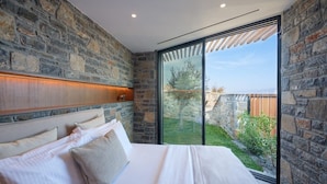 A bedroom with a stone wall, a large bed with white bedding, and a view of a garden through a glass door. - Villa Narethea, Divine Sea Views, Private Pool, Furnished Terrace, Crete (Crete)