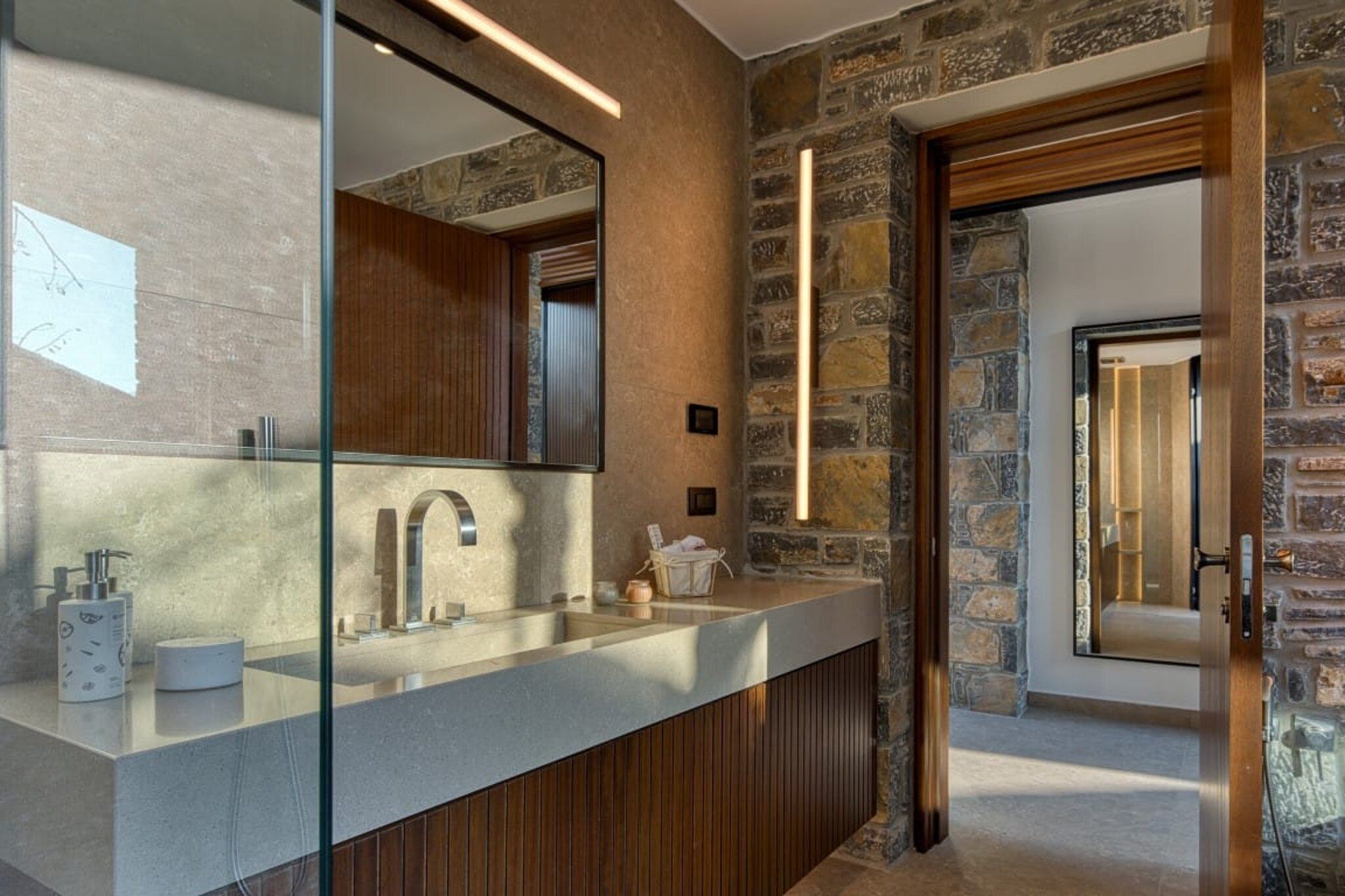 Een moderne badkamer met een stenen muur, een grote spiegel, een houten wastafelmeubel en een douchecabine met glazen wanden.