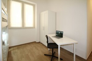A small, bright room with a white desk, a black chair, and a laptop. There is a window with blinds, a white wardrobe, and a staircase to the left.