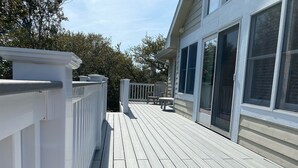 A wooden deck with a white railing, a seating area with chairs, and a building with large windows. - Escape to beautiful 4 BR home, Brightside OBX—Your Corolla Beach Retreat Awaits! (Corolla)