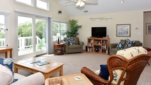 A spacious living room with a large window, a ceiling fan, a television, and a bookshelf. - Escape to beautiful 4 BR home, Brightside OBX—Your Corolla Beach Retreat Awaits! (Corolla)