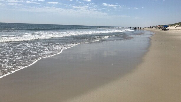 A wide sandy beach with gentle waves, a clear sky, and a few people in the distance. - Escape to beautiful 4 BR home, Brightside OBX—Your Corolla Beach Retreat Awaits! (Corolla)