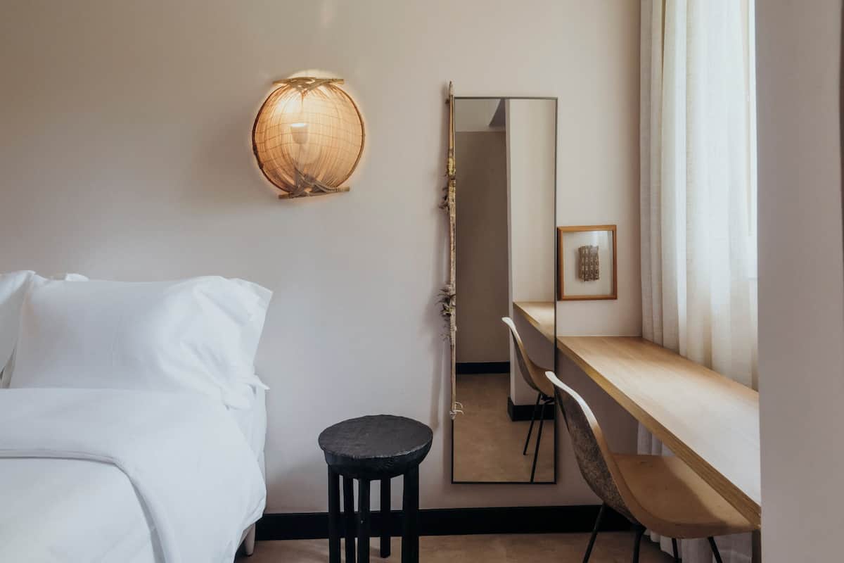 Ein Hotelzimmer mit einem Bett, einem kleinen Hocker, einem Schreibtisch, einem Spiegel und einer Wandlampe.