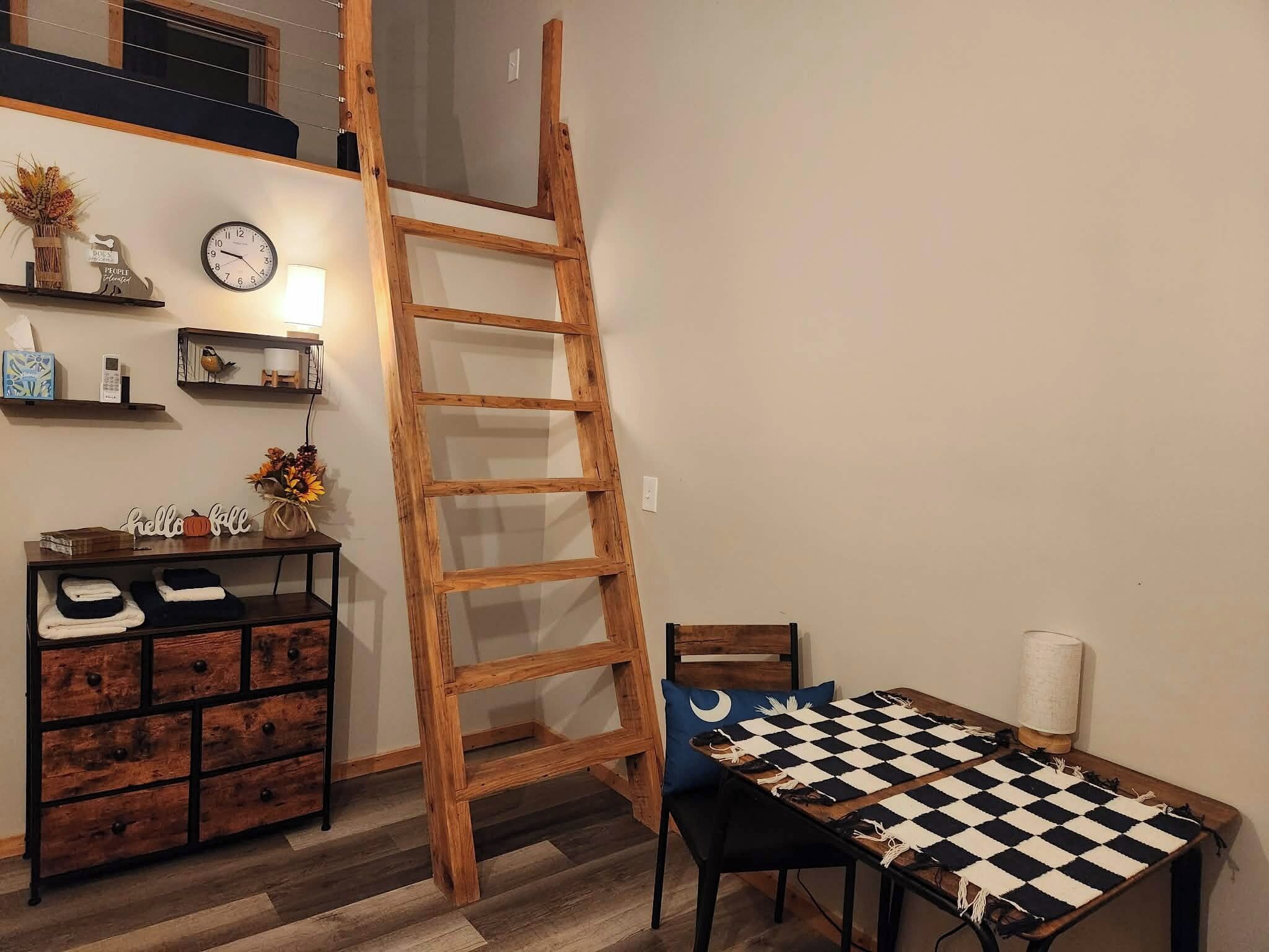 A room with a wooden ladder, a checkered table, a wooden cabinet, and a wall clock.