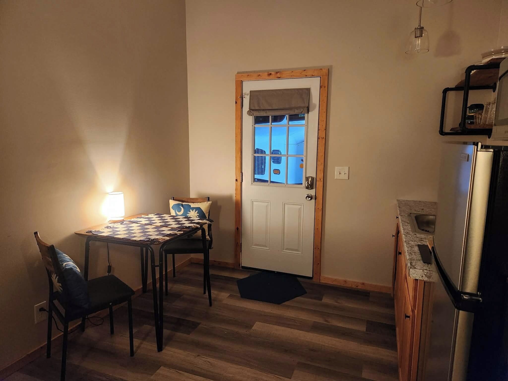 Une petite pièce chaleureuse avec une porte en bois, une table à carreaux, deux chaises, une lampe et un réfrigérateur.
