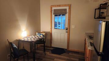 Une petite pièce chaleureuse avec une porte en bois, une table à carreaux, deux chaises, une lampe et un réfrigérateur.