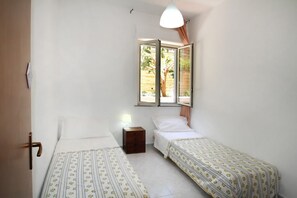 A small, well-lit room with two single beds, a small bedside table, and a window with a view of greenery outside.