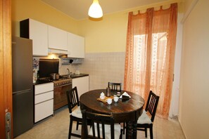Cocina compacta con armarios blancos, estufa y refrigerador. Hay una mesa redonda de madera con sillas y una ventana con cortinas.
