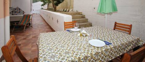 Un comedor con una mesa preparada para dos, sillas, una sombrilla verde y una escalera al fondo.