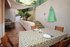 Zona pranzo con tavolo apparecchiato per due, sedie, un ombrello verde e una scala sullo sfondo.