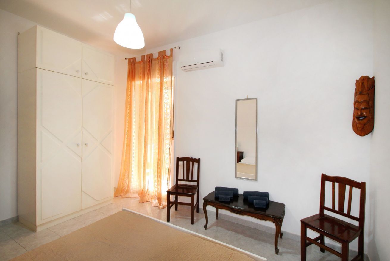 A room with a wardrobe, two chairs, a small table, a mirror, and a wooden mask on the wall.