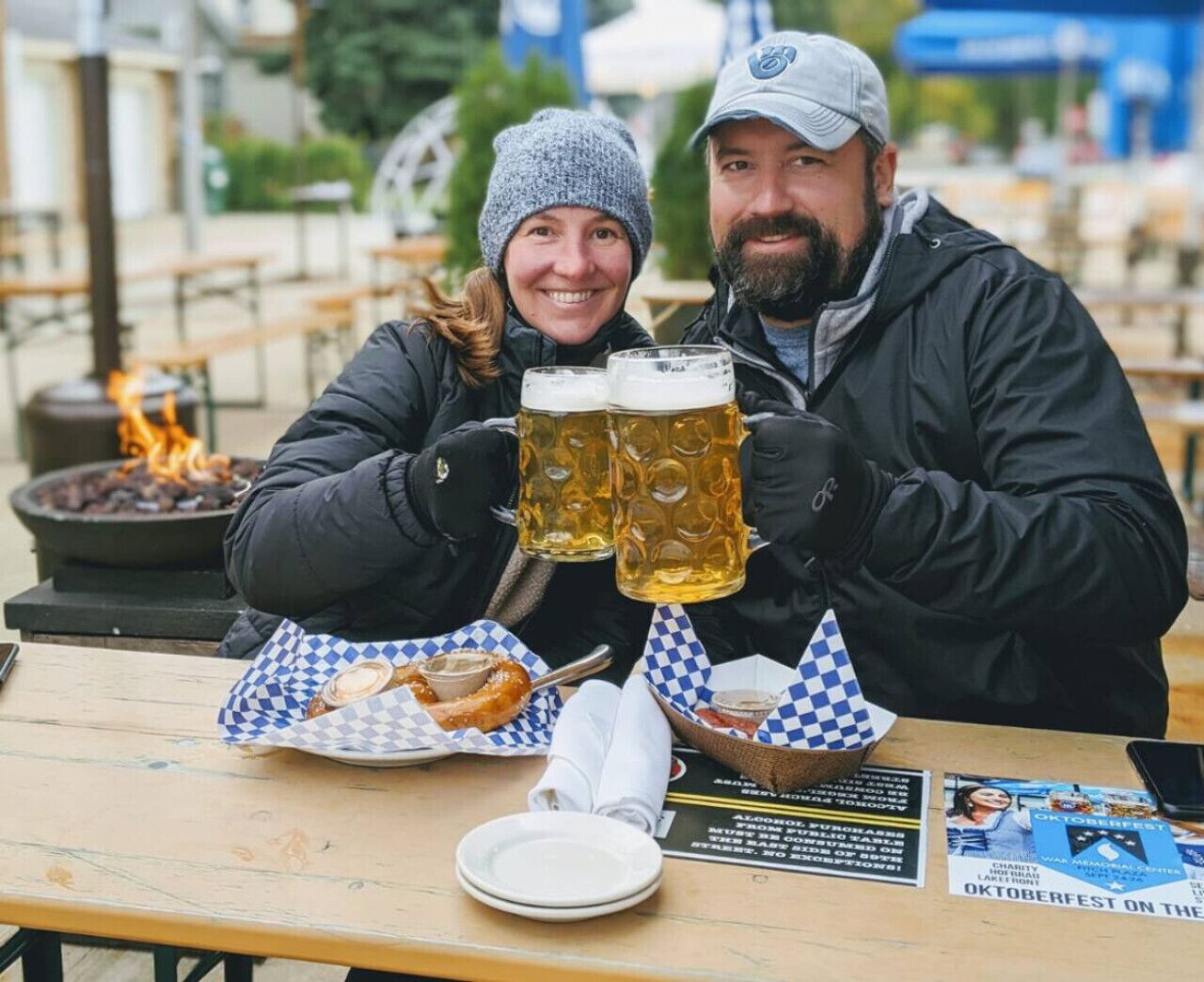 Un couple est assis à une table en plein air, devant des chopes de bière, une assiette de nourriture et une affiche annonçant l'Oktoberfest.