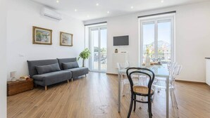 A modern living room with a grey sofa, a dining table with chairs, and large windows offering a view of the city.