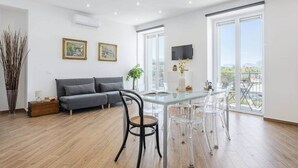 A modern living room with a grey sofa, a glass dining table, and clear chairs. There are two framed pictures on the wall, a potted plant, and a view of the outdoors through large windows.
