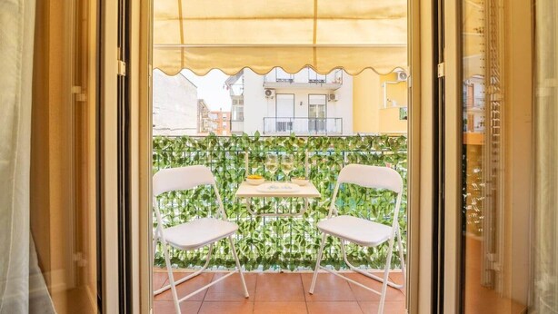 A balcony with two white chairs and a table, a yellow awning, and a green leafy wall. - nonMama | 2-Bath Stylish Apt near Mondello Beach (Palermo)
