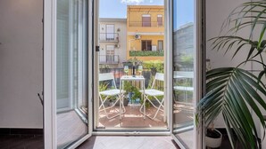 An open sliding glass door leading to a balcony with a table and chairs, potted plants, and a building in the background. - Mama | Your Stylish Oasis near Mondello Beach (Palermo)