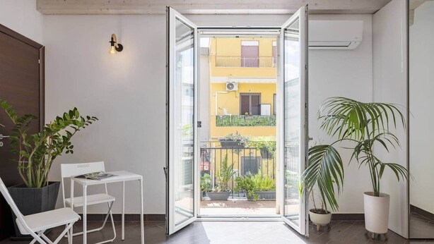 A modern apartment with a sliding glass door leading to a balcony with potted plants and a yellow building in the background. - Mama | Your Stylish Oasis near Mondello Beach (Palermo)