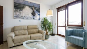 Un salon avec un canapé beige, un canapé bleu, une table basse en verre, une plante en pot, un grand tableau représentant un phare et une fenêtre avec des stores.
