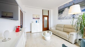 Un salon moderne avec un canapé beige, une table basse en verre, un téléviseur à écran plat et un tableau représentant un phare.