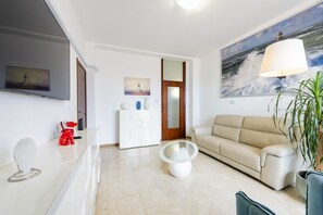 A modern living room with a beige sofa, a glass coffee table, a flat-screen TV, and a painting of a lighthouse.