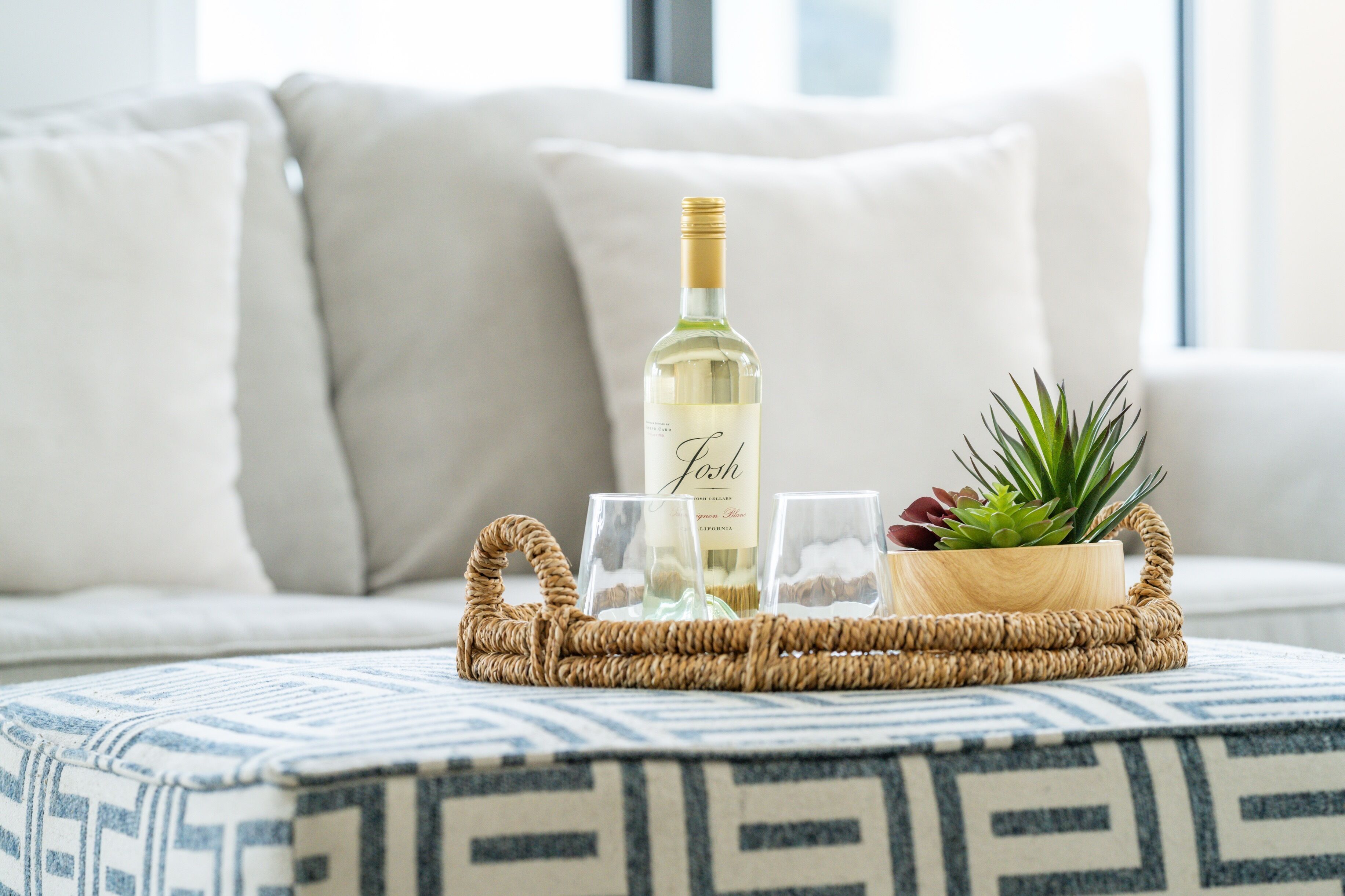 Een fles wijn, twee glazen en een kleine plant op een geweven dienblad op een gestreept kussen.